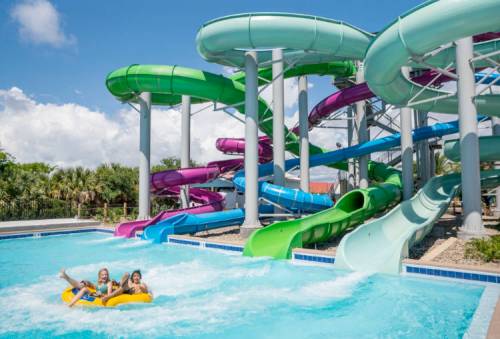Three children ride a yellow inflatable in a pool, with colorful water slides in the background under a blue sky.