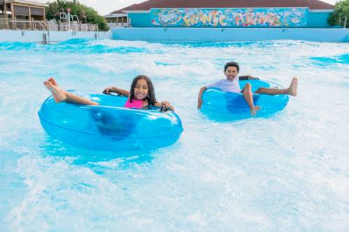 Two children float on blue inner tubes in a wave pool, enjoying a sunny day at a water park.