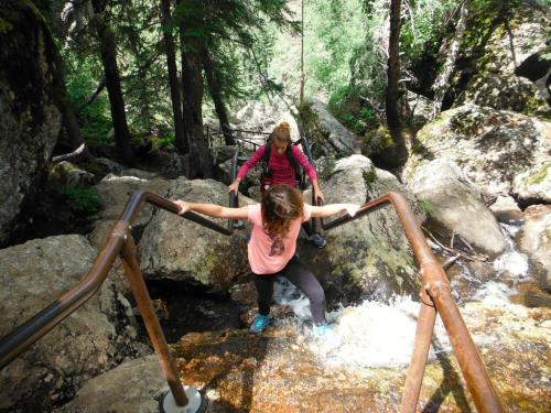Two hikers navigate rocky terrain with a stream, using handrails for support in a lush, green forest.