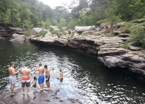 A group of people swimming and relaxing by a rocky swimming hole surrounded by trees.