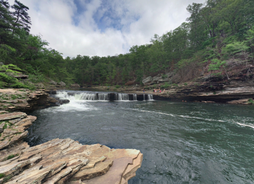 A serene river flows over rocks, surrounded by lush green trees and a cloudy sky.