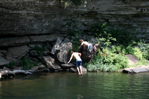 Two boys jumping off a rock into a calm, green river surrounded by lush vegetation and a rocky cliff.