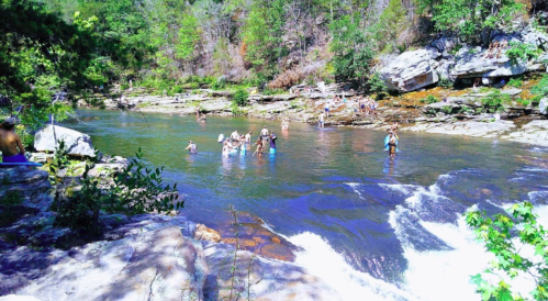 People enjoying a sunny day by a river, swimming and relaxing among rocks and greenery.