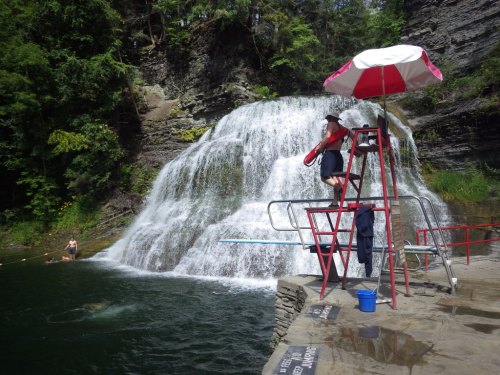 A lifeguard on a tall chair watches over a waterfall and swimming area, with trees surrounding the scene.