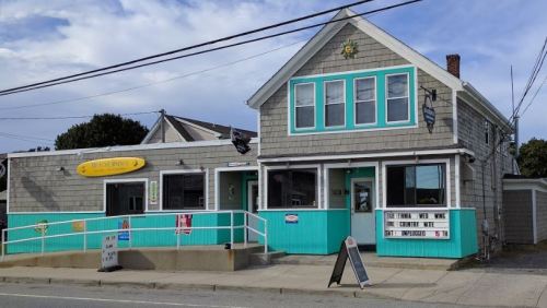 A charming coastal restaurant with a turquoise exterior and large windows, featuring outdoor seating and a welcoming sign.