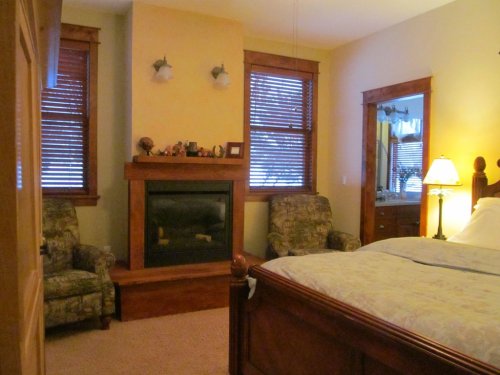 Cozy bedroom with a fireplace, two armchairs, a mirror, and warm lighting from a bedside lamp.