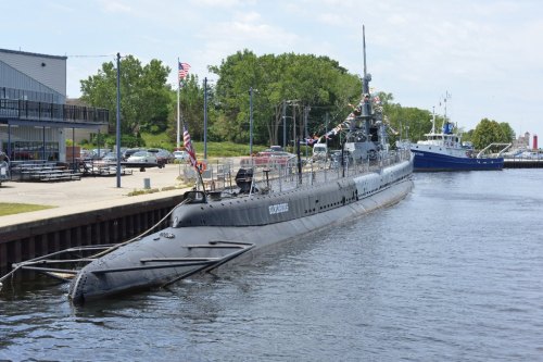 A submarine docked at a waterfront, decorated with flags, alongside a boat and greenery in the background.