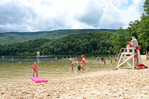 A sandy beach with people swimming and playing in the water, surrounded by green hills and a lifeguard on duty.