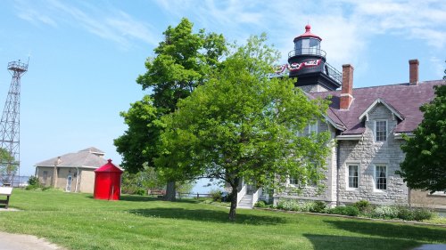 A lighthouse beside a green lawn, trees, and a red shed, with a clear blue sky in the background.