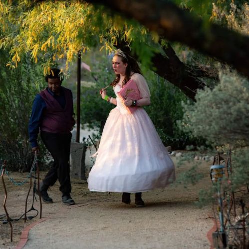 A woman in a pink ball gown and tiara walks along a garden path, while a man in a purple vest follows her.