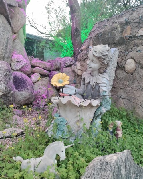 A whimsical garden scene featuring a fairy statue with a flower, surrounded by rocks and greenery.