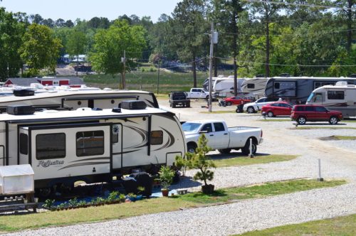 A gravel RV park with several parked RVs and vehicles, surrounded by trees and open space.