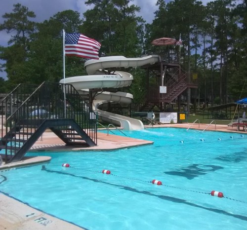 A swimming pool with a water slide, American flag, and lane markers, surrounded by trees and a cloudy sky.