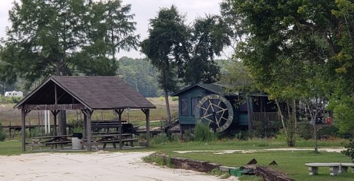 A rustic scene featuring a gazebo and a green building with a water wheel, surrounded by trees and open land.