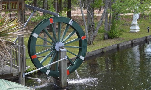 A green water wheel beside a calm pond, with trees and a statue in the background.