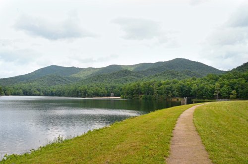 A serene lake surrounded by green mountains, with a walking path along the water's edge under a cloudy sky.