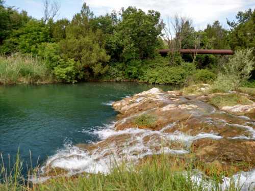 A serene pond surrounded by lush greenery and rocky edges, with gentle water flowing over the rocks.