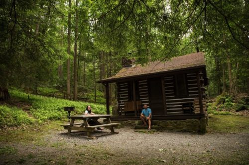 A rustic cabin in a forested area, with two people sitting at a picnic table nearby.