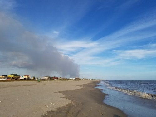 A sandy beach stretches along the shore, with a blue sky and distant smoke rising from the land.