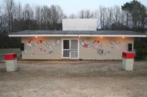 A small building with colorful board game murals on the exterior, surrounded by trees and gravel.