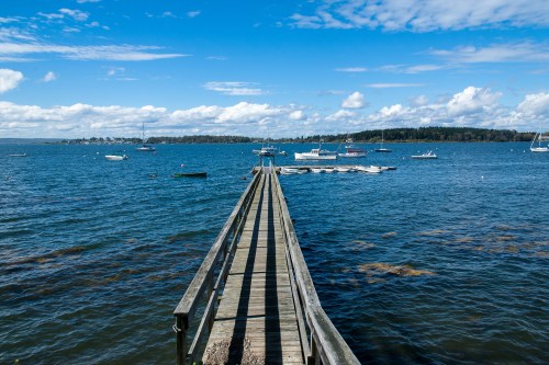 A wooden pier extends into a calm blue sea, with boats floating and a clear sky dotted with clouds.