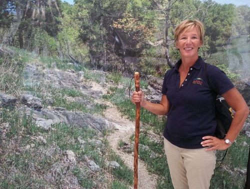 A woman stands with a walking stick in front of a scenic nature backdrop, smiling and wearing a polo shirt.