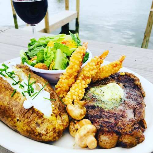 A plate featuring a baked potato, grilled steak, shrimp, salad, and a glass of red wine by the water.