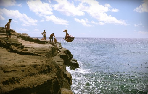 A person jumps off a rocky cliff into the ocean, while others watch from the shore under a sunny sky.