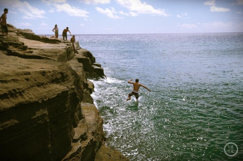 A person jumps off a rocky ledge into the ocean, with others nearby enjoying the beach on a sunny day.