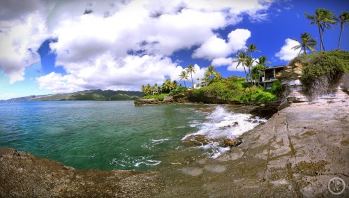 A scenic coastal view with rocky shores, palm trees, and vibrant blue water under a partly cloudy sky.