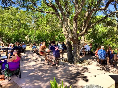 Outdoor dining area with people seated at tables under trees, enjoying a sunny day in a lush garden setting.