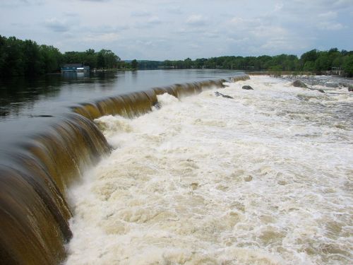 A wide river with a cascading waterfall, surrounded by green trees and a cloudy sky.