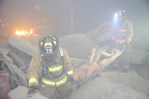 Firefighters in protective gear rescue a person on a stretcher from a smoky, debris-filled environment.