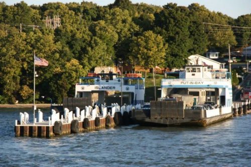 Two ferries docked at a pier, surrounded by trees and a small building in the background, with an American flag nearby.