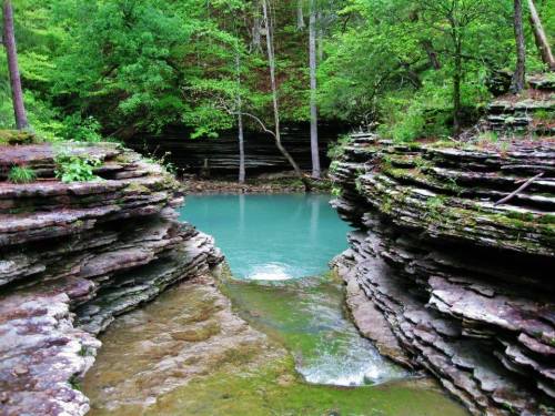 A serene turquoise pool surrounded by layered rock formations and lush green trees in a tranquil natural setting.