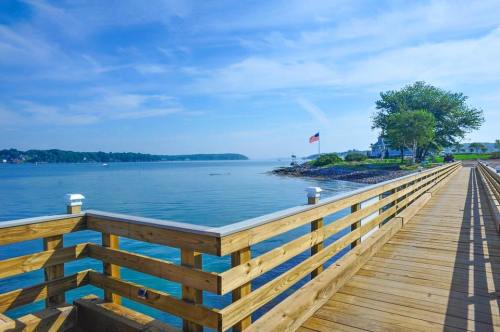 A wooden pier extends over calm water, with trees and a flag in the background under a clear blue sky.