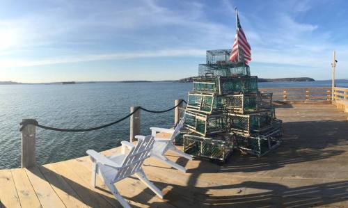 A wooden dock by the water features a stack of lobster traps and two white chairs, with an American flag waving above.
