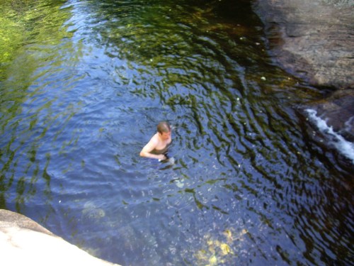 A person swimming in a clear, calm body of water surrounded by rocks and greenery.