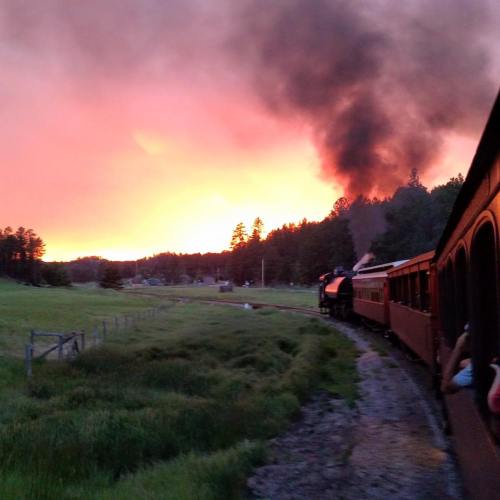 A vintage steam train travels through a scenic landscape at sunset, with smoke billowing against a colorful sky.