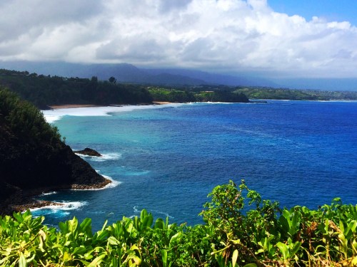 Scenic coastal view with turquoise waters, rocky cliffs, and lush greenery under a partly cloudy sky.