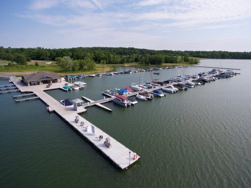 Aerial view of a marina with multiple boats docked along a long wooden pier, surrounded by green trees and calm water.