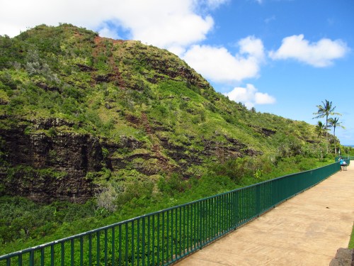 A scenic pathway along a lush green hillside under a blue sky with scattered clouds.