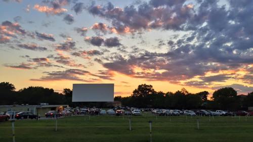 A drive-in theater at sunset, with cars parked in front of a large screen and colorful clouds in the sky.
