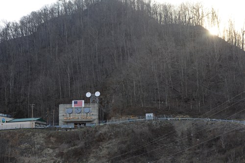 A small building with an American flag, set against a wooded hillside at sunset.