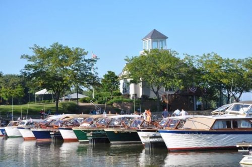 A marina with several boats docked, surrounded by trees and a building in the background under a clear blue sky.