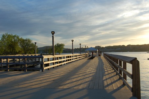 A wooden pier extends into a calm body of water, with lampposts casting shadows under a colorful sky at sunset.