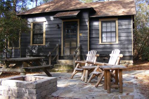 A cozy cabin with a wooden porch, picnic table, and two chairs near a stone fire pit in a wooded area.