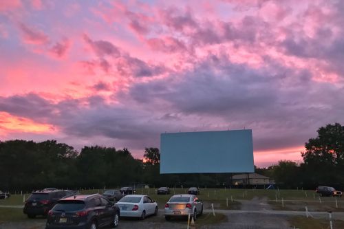 A drive-in movie theater with an empty screen at sunset, surrounded by parked cars and colorful clouds.