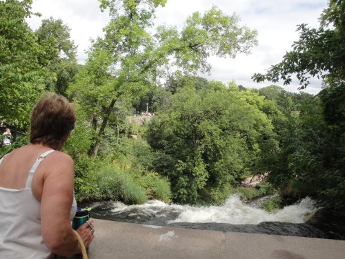 A person sits by a waterfall surrounded by lush green trees and a scenic view of nature.