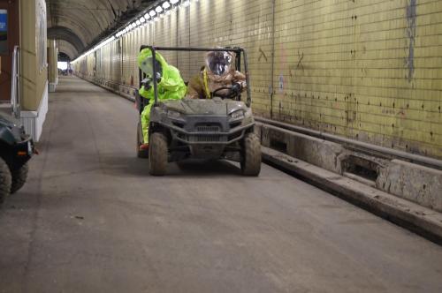 Two people in protective gear drive a utility vehicle down a long, dimly lit tunnel with yellow walls.
